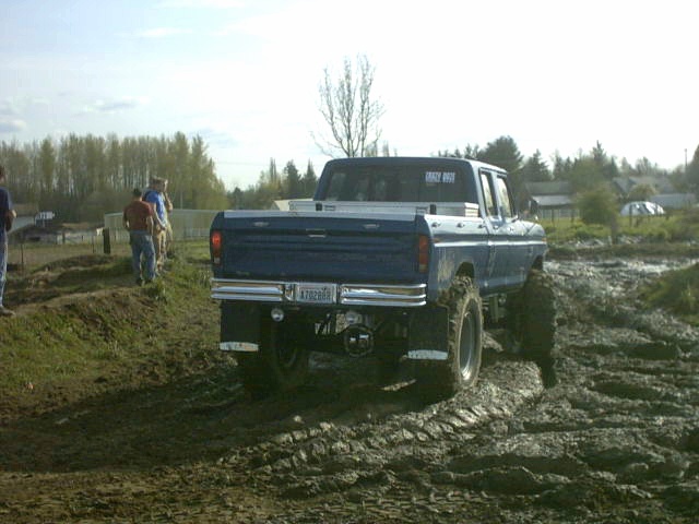 Blue lifted Ford crew-cab pickup with bed toolbox seen from the rear at a muddy off-road event