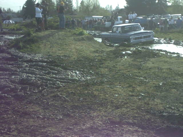 White and blue vintage Ford pickup at a muddy mud bog course with spectators