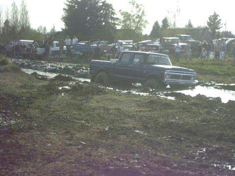 1995 Ford Bronco - A dark blue Ford pickup truck navigates a muddy track with spectators gathered nearby.