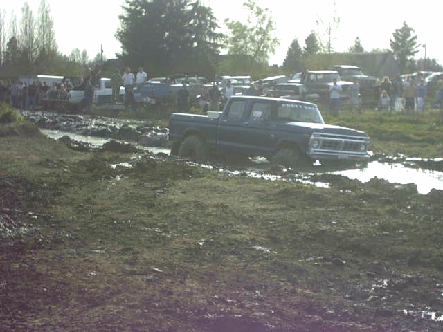 Dark blue 1970s Ford crew-cab pickup stuck in a muddy field at a mud-bog event