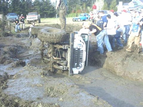 1995 Ford Bronco - An overturned white SUV is stuck in thick mud, surrounded by onlookers.