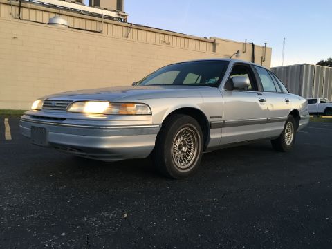 1994 Ford Crown Victoria - A silver 1994 Ford Crown Victoria is parked in a lot with a building in the background.