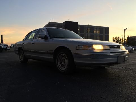 1994 Ford Crown Victoria - Silver sedan parked outside a building with the headlights on, under a dusky sky.