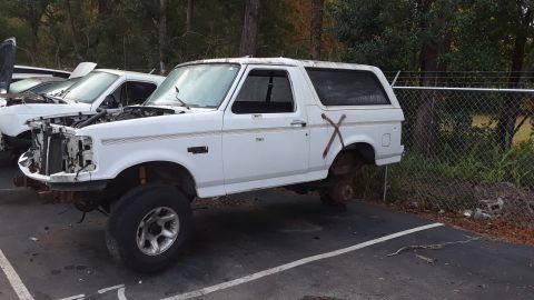 1994 Ford Bronco - White Ford Bronco missing wheels and front bumper, parked in a lot with another vehicle.