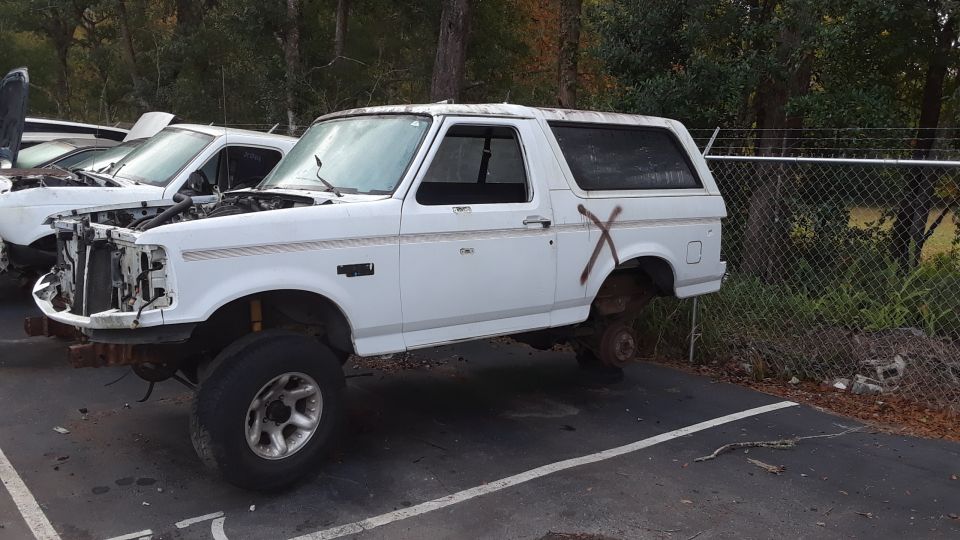 White Ford Bronco missing front bumper and rear wheels, parked next to another vehicle with hood open