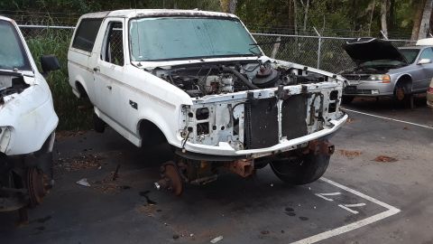 1994 Ford Bronco - A white Ford Bronco is stripped of its front bumper, grille, and headlights in a junkyard setting.