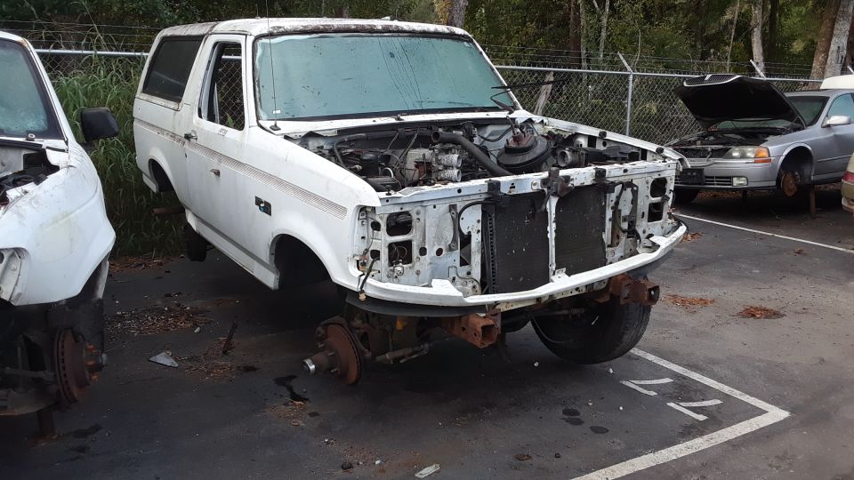 White Ford Bronco missing front end parts, sitting in a junkyard next to another vehicle