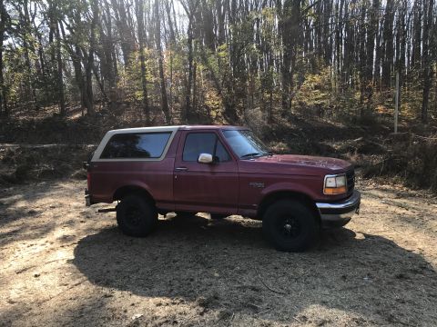 1994 Ford Bronco - A maroon Ford Bronco is parked on a dirt path surrounded by trees.
