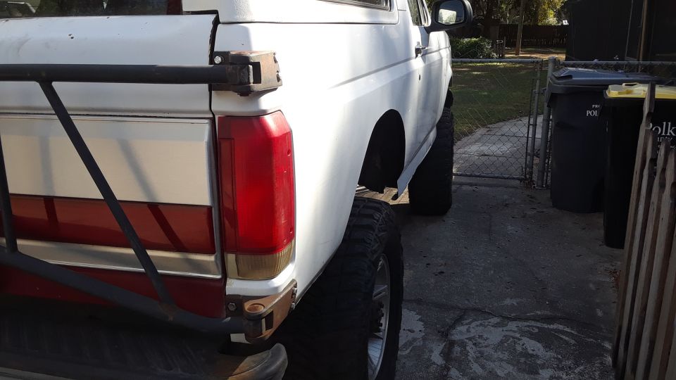 White Ford Bronco with red stripe, lifted suspension, and aftermarket rear bumper. Visible from the rear quarter panel