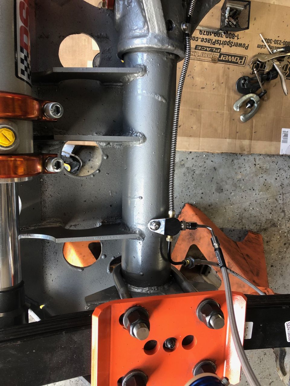 Close-up of a vehicle suspension component, possibly a shock absorber mount, with a brake line attached. Tools and cardboard are visible in the background