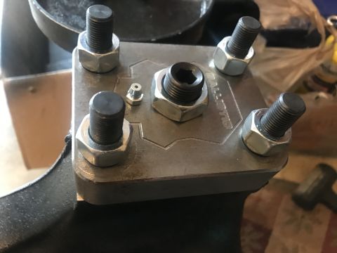 1989 Ford Bronco - A close-up view of a metal bracket with four bolts and nuts, a central hex bolt, and a small grease fitting. The bracket has the word "BALL…