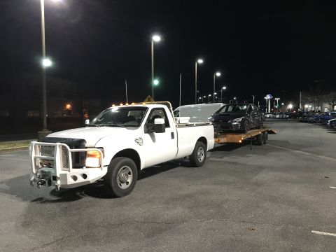 2008 Ford F-250 - A white Ford F-250 pickup truck tows a flatbed trailer carrying a dark-colored sedan at night in a parking lot.