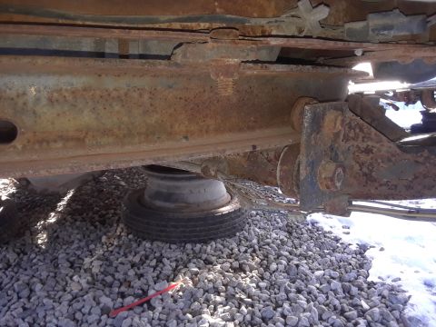 1994 Ford Bronco - The underside of a vehicle frame shows significant rust on the metal beams and brackets. A spare tire is visible resting on gravel below th…