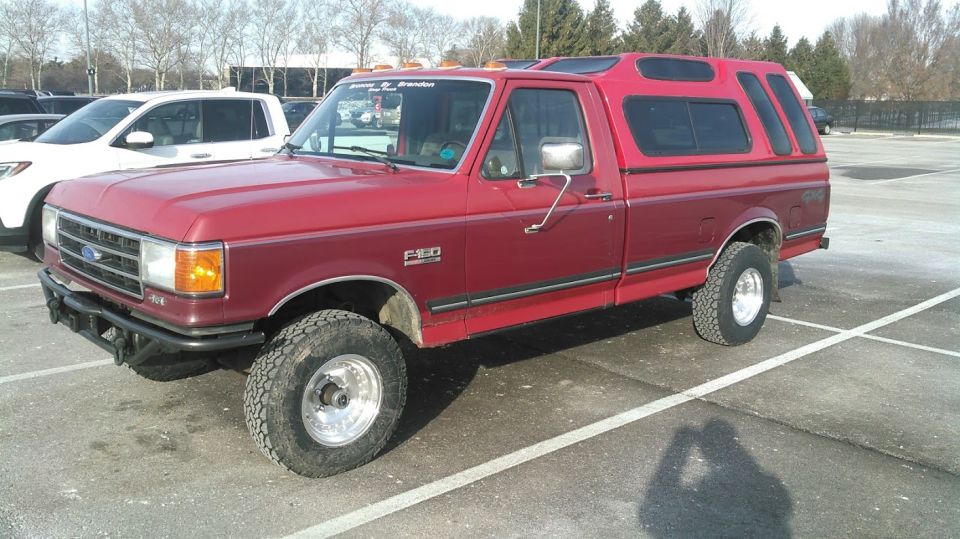 Red 1991 Ford F-150 pickup truck with camper shell and custom bumper in a parking lot.