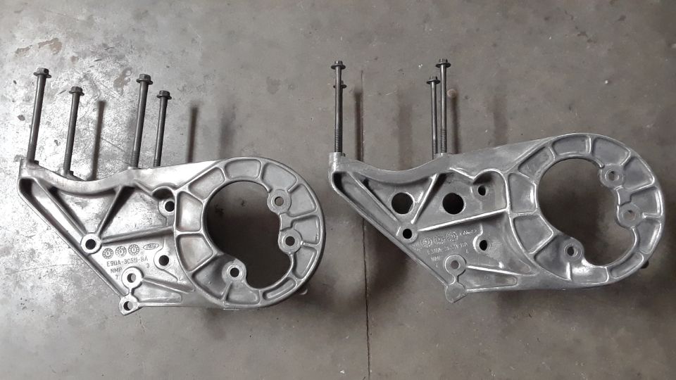Two silver aluminum engine brackets with bolts on a concrete surface