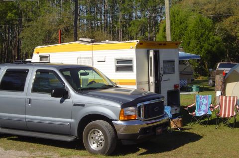 1974 Ford Bronco - A silver SUV is parked in front of a travel trailer, with camping chairs and a tent visible in the background.