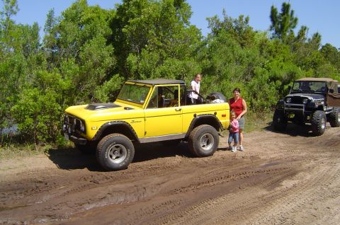 1974 Ford Bronco - A yellow 1974 Ford Bronco is parked on a dirt road with two women and two children nearby, while another vehicle is visible in the backgrou…