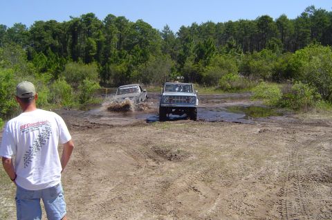 1974 Ford Bronco - Two vehicles navigate a muddy off-road course with trees in the background.