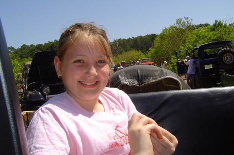 1974 Ford Bronco - A young girl smiles while sitting in an off-road vehicle, with a gathering of people and other vehicles visible in the background on a sunn…