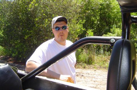 1974 Ford Bronco - Man in sunglasses and cap leans on vehicle interior with trees behind him.