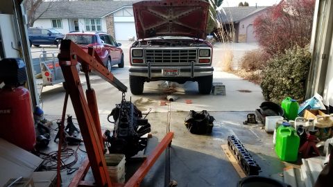 1985 Ford Bronco - A red Ford truck is undergoing engine repair, with the engine hoisted out and parts laid out on the ground.