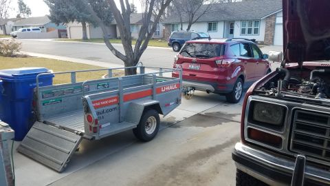 1985 Ford Bronco - A red Ford Escape is hitched to a U-Haul trailer in a driveway, with an older red truck's engine exposed in view.