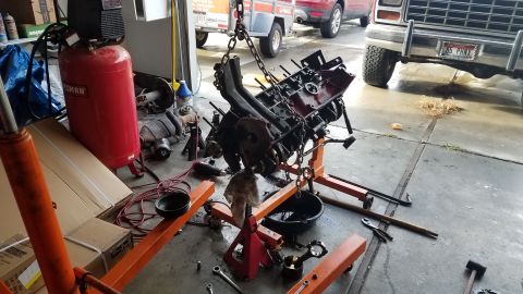 1985 Ford Bronco - An engine is suspended by chains over an engine stand, with various tools and a red air compressor nearby.