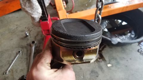 1985 Ford Bronco - A mechanic holds a used engine piston, likely removed during repair work, with tools and an oil pan visible.