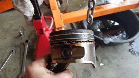 1985 Ford Bronco - A piston and connecting rod are held up for inspection, with engine repair equipment visible.