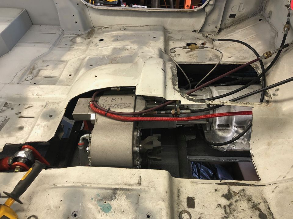 White vehicle interior floor pan with exposed drivetrain, wiring, and hydraulic lines. A silver electric motor is visible