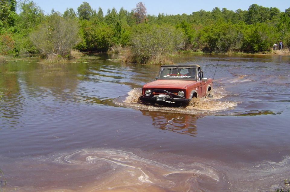 Red early Ford Bronco with winch fording deep brown water past pine trees