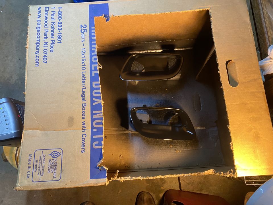 Two black car door handles inside a cardboard box, one is partially painted