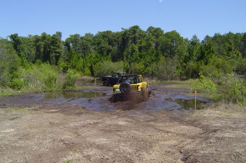 1974 Ford Bronco - Two off-road vehicles navigate a muddy track with trees in the background.
