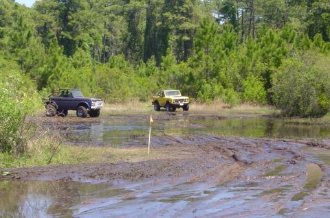 1974 Ford Bronco - Two Ford Broncos navigate a muddy off-road course surrounded by trees.