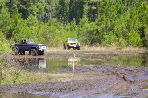 1974 Ford Bronco - Two off-road vehicles navigate a muddy track surrounded by trees.