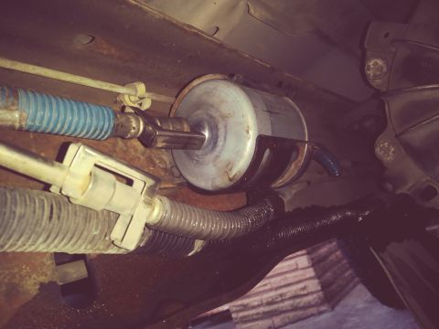 1996 Ford Bronco - Cylindrical inline fuel filter mounted to the underbody, with rusty fuel and brake lines and a blue vacuum hose nearby.