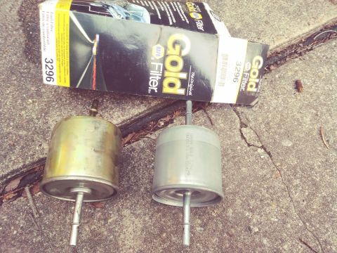 1996 Ford Bronco - Old and new cylindrical inline fuel filters lying on pavement next to a black and gold Motorcraft Gold 3296 filter box.