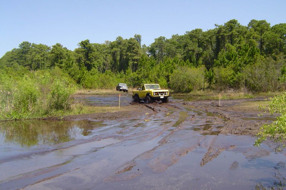 Yellow open-top early Ford Bronco crossing a rutted mud pit at an off-road park
