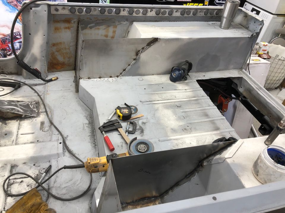 Interior of a vehicle being worked on, showing metal floor and body panels, with tools like a welder and grinder present