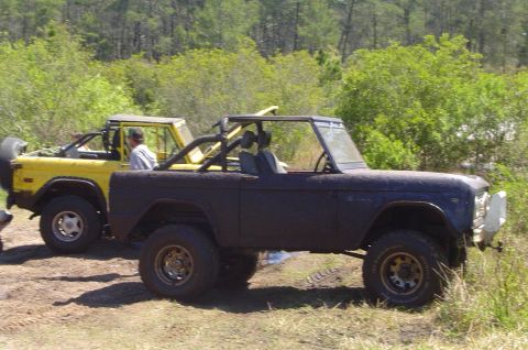 1974 Ford Bronco - Two 1974 Ford Broncos are seen off-roading on a sunny day with trees and brush surrounding them.