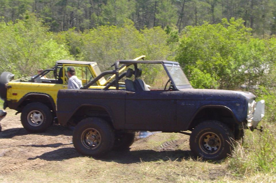 Muddy blue doorless early Ford Bronco with roll cage beside a yellow Bronco