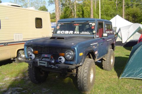 1974 Ford Bronco - A blue Ford Bronco is parked on grass next to an RV and several tents, suggesting a campsite setting.