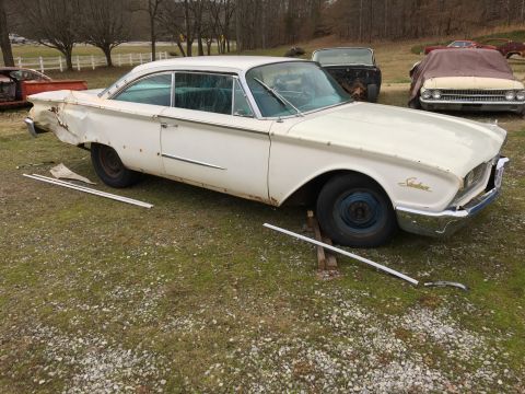 1960 Ford Starliner - A white 1960 Ford Starliner sits in a yard, showing damage to its rear and other vehicles in the background.