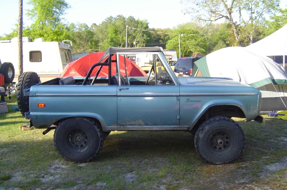 Light blue early Ford Bronco side profile with roll cage and black steel wheels at a campground