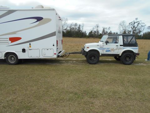 1992 Ford Bronco - A white off-road vehicle tows a large RV across a grassy field under a cloudy sky.