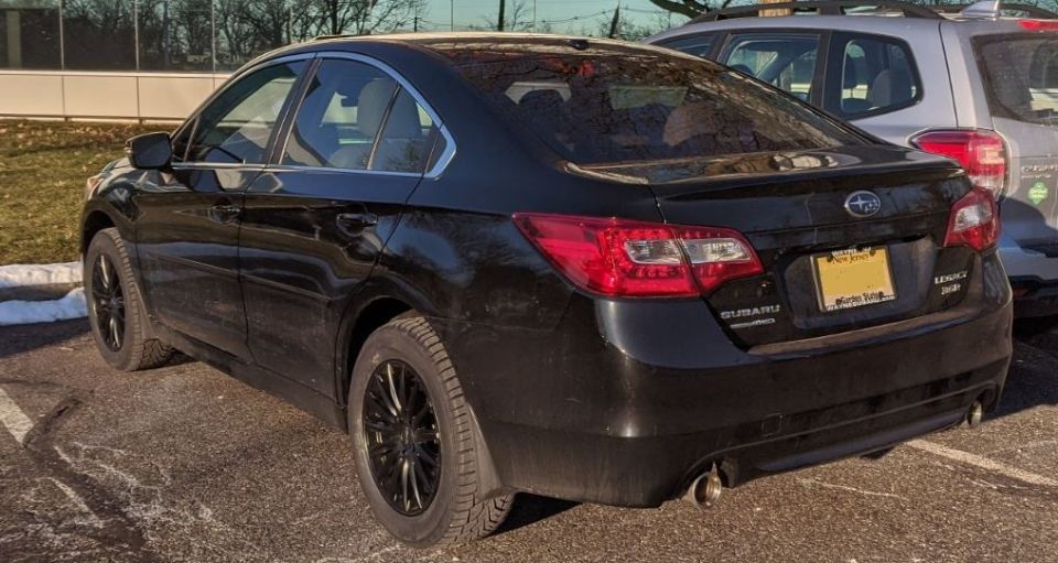 Black Subaru Legacy sedan with black wheels, rear three-quarter view in a parking lot