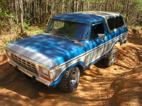 1978 Ford Bronco - Elevated 3/4 front view of a weathered blue and white late-'70s Ford Bronco on a red dirt road in pine woods, with faded oxidized paint, ru…