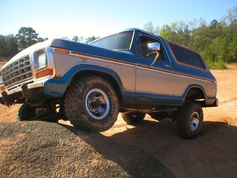 1978 Ford Bronco - Blue and silver two-tone full-size Ford Bronco flexing on a rock ledge at a red-clay off-road park, front driver wheel lifted to show the s…