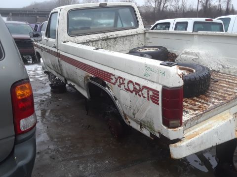 1994 Ford Bronco - The rear passenger side of a white Ford pickup truck is shown in a junkyard setting. The truck bed contains two tires, and the side of the…