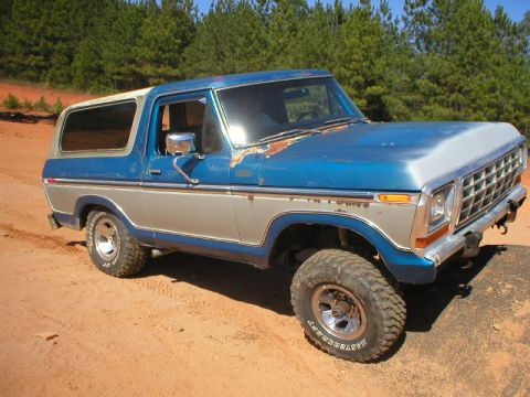 1978 Ford Bronco - A two-tone 1978 Ford Bronco with off-road tires sits on a dirt track, surrounded by trees.
